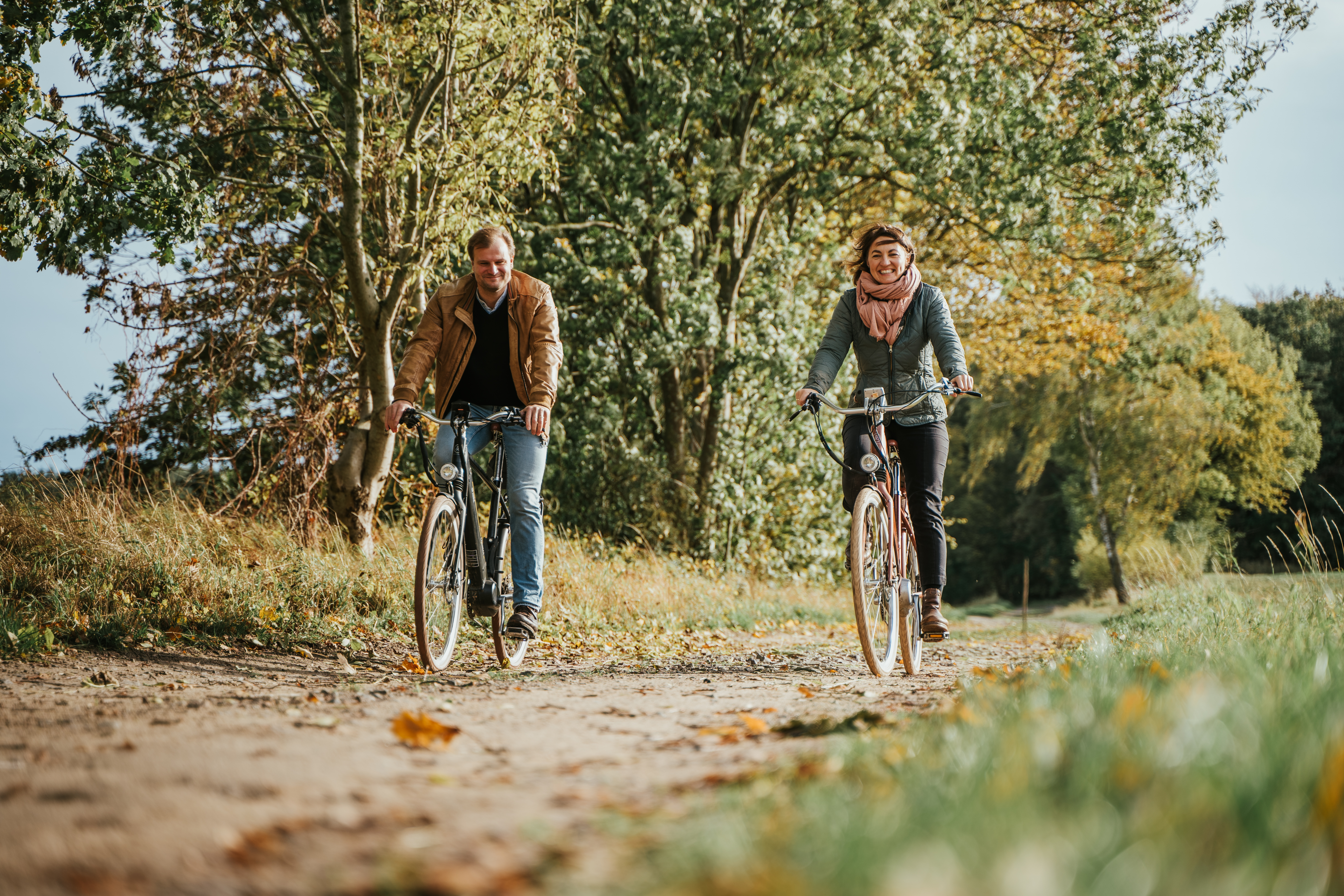 Ein Mann und eine Frau fahren auf Fahrrädern über einen Feldweg. Auf dem Boden liegen Blätter und die Bäume sind schon leicht herbstlich gefärbt